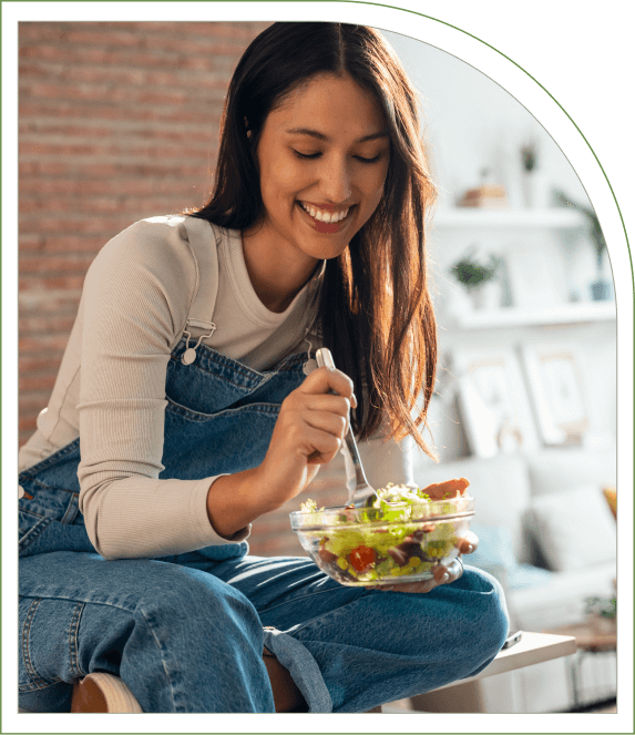 A woman in overalls sits on a counter, smiling while eating a salad in a modern, well-lit kitchen.