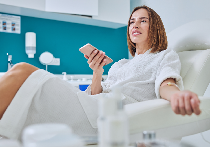 A woman in a white robe sits in a chair, holding a smartphone with a relaxed expression. The room has teal walls and various beauty products are visible in the foreground.