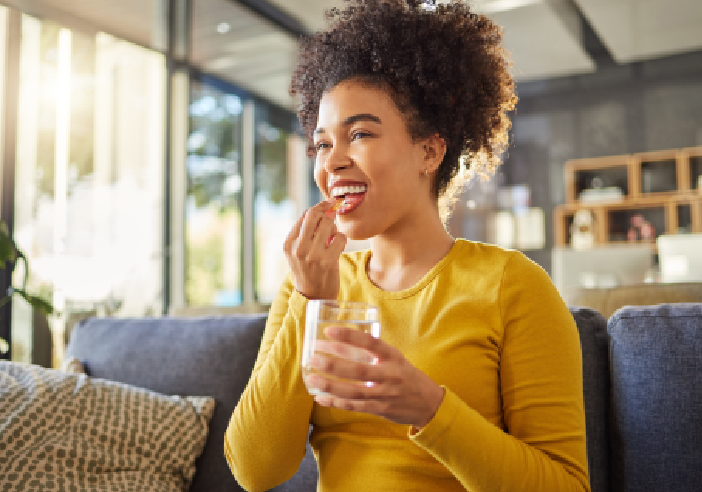 A person in a yellow shirt is sitting on a couch, holding a glass of water and taking a pill, in a bright, modern living room.