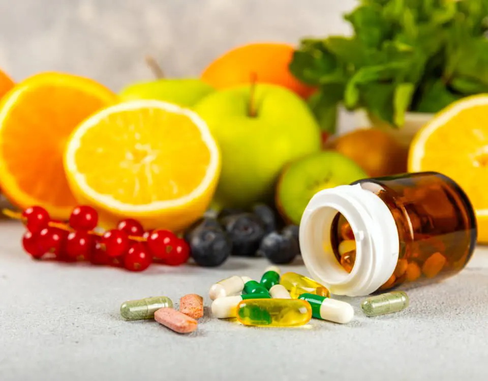 Open pill bottle with assorted capsules and tablets on a surface, surrounded by fresh fruits like oranges, apples, and berries.