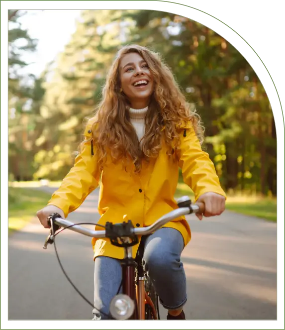 A person with long curly hair and a yellow jacket is happily riding a bicycle on a sunny day in a park.