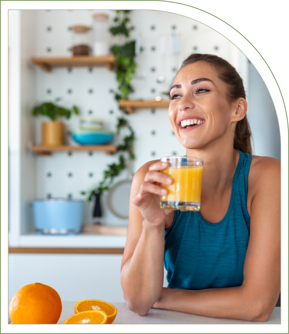 A woman in a blue tank top smiles while holding a glass of orange juice in a kitchen with shelves in the background. Two oranges are on the counter.