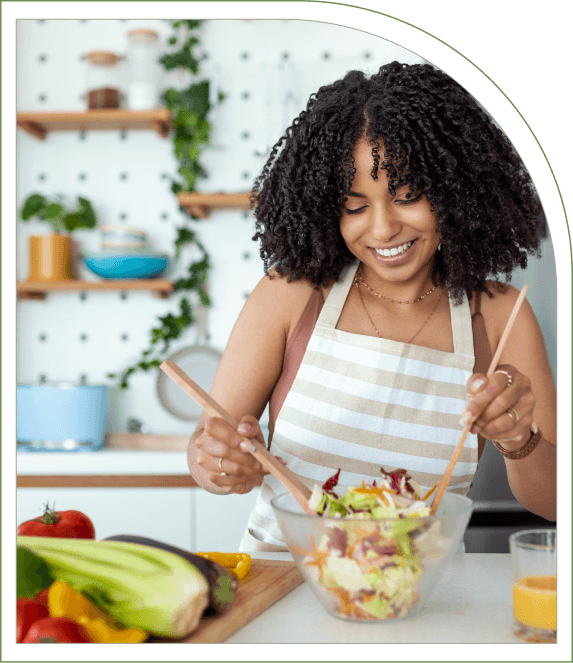 A woman in a striped apron is smiling while mixing a salad in a glass bowl in a kitchen with plants and vegetables around.