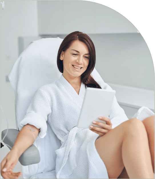 A woman in a white robe sits in a chair, smiling while reading a tablet.