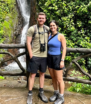 Two people stand in front of a waterfall, posing for a photo on a wooden bridge surrounded by greenery. Both wear casual outdoor clothing and hiking boots.